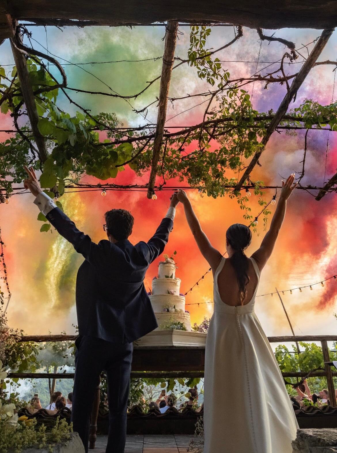 Coppia di sposi che alza le braccia di fronte alla torta mentre il cielo esplode di fumi multicolore.